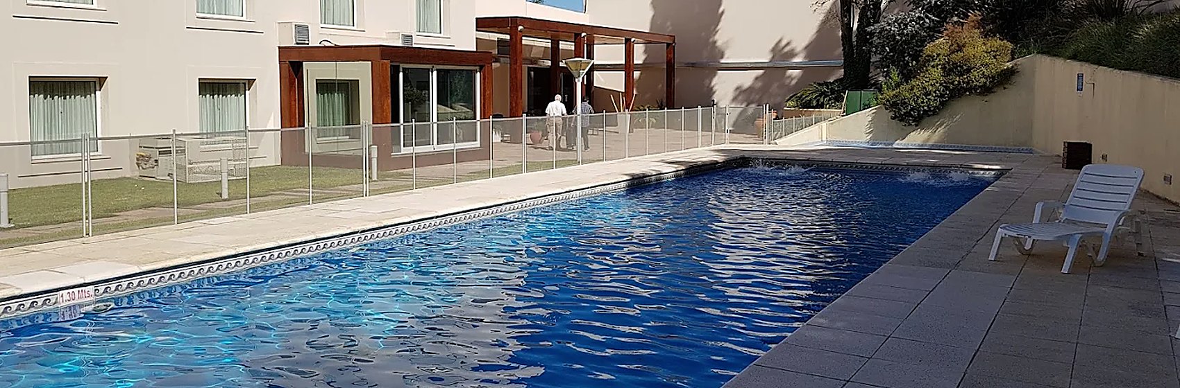 Pileta exterior junto a un edificio blanco de tres niveles, con cipreses y cielo despejado.