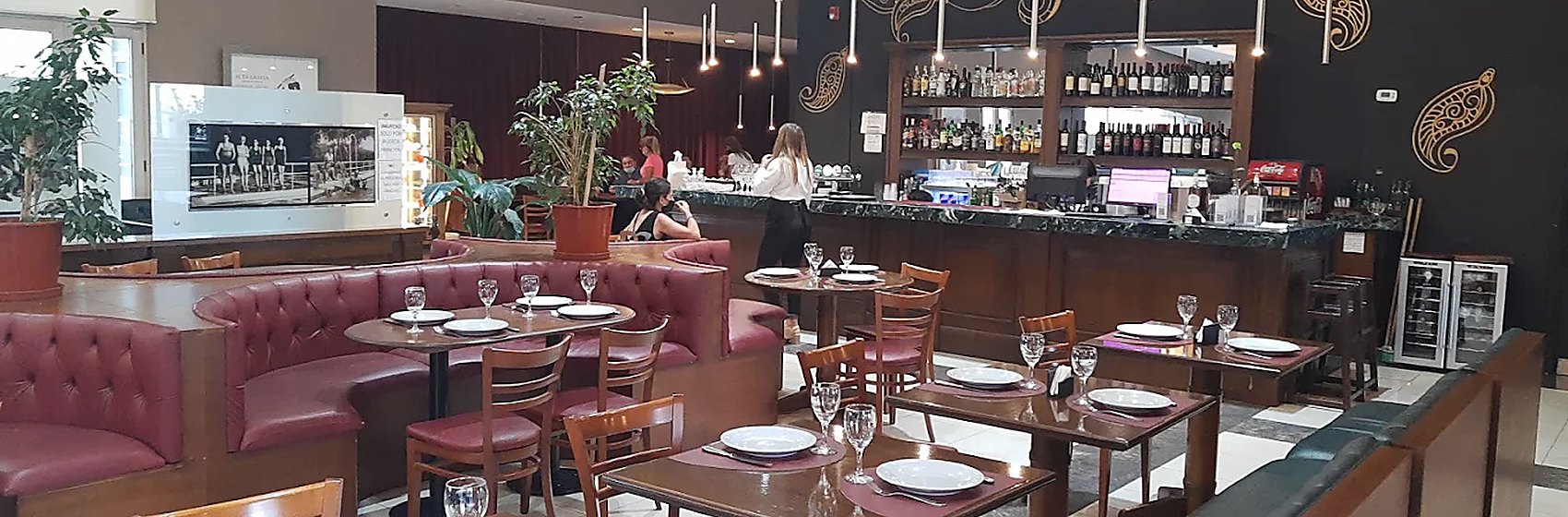 Comedor de hotel con banquetas tapizadas, mesas servidas y barra al fondo.